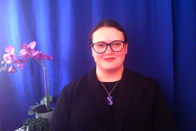 Photo of white woman with dark hair and dark glasses, wearing a black jacket with a necklace. The background is a dark blue with plants on a small table, behind and to the right or her.